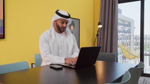 Completing projects and tasks at home, enjoying hot drinks, a picture of a young Emirati Gulf Arab man wearing a white kandura and ghutra sitting on a gray sofa, using a laptop, conducting work discussions, having a good time, the concept of technology and using modern technical devices