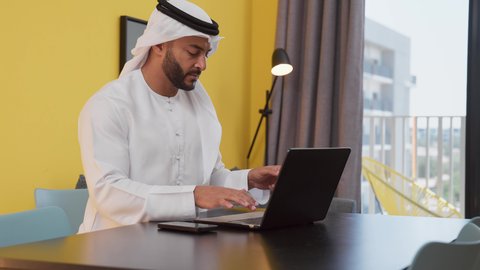 A photo of a young Emirati Gulf Arab man wearing a white kandura and ghutra sitting on a gray sofa, using a laptop, conducting work discussions, having a good time, the concept of technology and using modern technical devices, completing projects and tasks at home, enjoying hot drinks