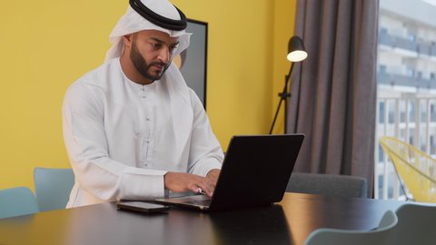 Conducting business discussions, having fun, the concept of technology and using modern technology devices, completing projects and tasks at home, enjoying hot drinks, a picture of a young Emirati Gulf Arab man wearing a white kandura and ghutra sitting on a gray sofa, using a laptop computer