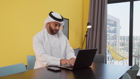 The concept of technology and the use of modern technology devices, completing projects and tasks at home, enjoying hot drinks, a picture of a young Emirati Gulf Arab man wearing a white kandura and ghutra sitting on a gray sofa, using a laptop, having a good time