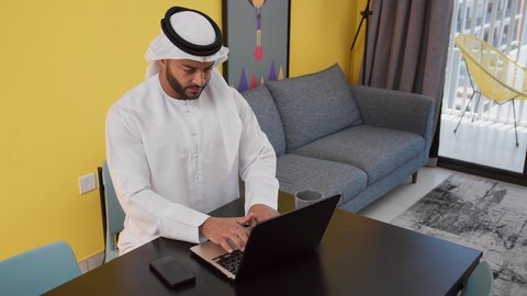 Completing projects and tasks at home, enjoying hot drinks, a picture of a young Emirati Gulf Arab man wearing a white kandura and ghutra sitting on a gray sofa, using a laptop, the concept of technology and the use of modern technical devices, having a good time