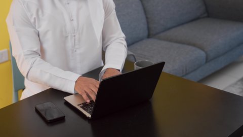 A photo of a young Emirati Gulf Arab man wearing a white kandura and ghutra sitting on a gray sofa, using a laptop, completing projects and tasks in a cafe, the concept of technology and the use of modern technical devices, having a good time, enjoying hot drinks