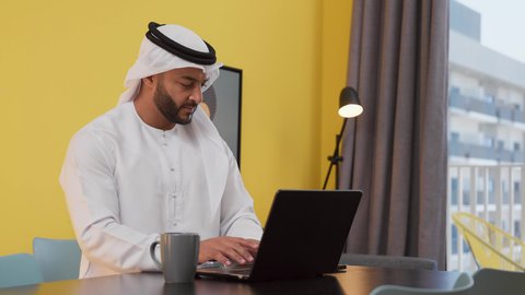 الاستمتاع بالمشروبات الساخنه ، تصوير لشاب عربي خليجي إماراتي يرتدي الكندورة والغترة البيضاء يجلس على اريكة رمادية ، يستخدم جهاز الحاسوب المحمول ، انجاز المشاريع والمهام في الكافيه ، مفهوم التكنولوجيا وإستخدام أجهزة تقنية حديثة ، قضاء وقت ممتع