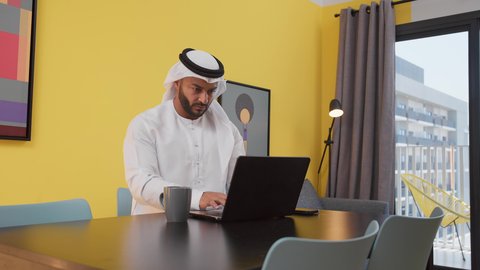 مفهوم التكنولوجيا وإستخدام أجهزة تقنية حديثة ، انجاز المشاريع والمهام في الكافيه ، قضاء وقت ممتع ,تصوير لشاب عربي خليجي إماراتي يرتدي الكندورة والغترة البيضاء يجلس على اريكة رمادية ، يستخدم جهاز الحاسوب المحمول ، الاستمتاع بالمشروبات الساخنه