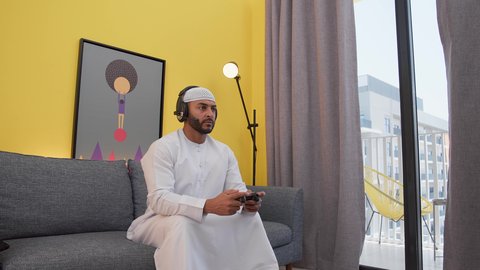 رجل عربي اماراتي خليجي يرتدي الكندورة والطاقيه وسماعات الرأس ، يمسك عصا التحكم ،تعابير الاندماج والتفاعل والتركيز، قضاء اوقات ممتعه ، دمج التقنيات الحديثه فى المتعة والتسلية ، ألعاب الفيديو الترفيهية