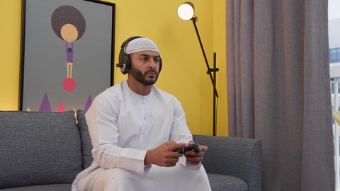تعابير الاندماج والتفاعل والتركيز، قضاء اوقات ممتعه ، دمج التقنيات الحديثه فى المتعة والتسلية ، رجل عربي اماراتي خليجي يرتدي الكندورة والطاقيه وسماعات الرأس ، يمسك عصا التحكم ، ألعاب الفيديو الترفيهية