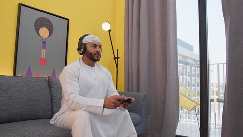 رفع قبضة اليد بتعابير الفوز ، دمج التقنيات الحديثه فى المتعة والتسلية ، قضاء اوقات ممتعه ، رجل عربي اماراتي خليجي يرتدي الكندورة والطاقيه وسماعات الرأس ، يمسك عصا التحكم ، ألعاب الفيديو الترفيهية