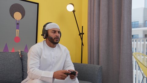 دمج التقنيات الحديثه فى المتعة والتسلية ، قضاء اوقات ممتعه ، رجل عربي اماراتي خليجي يرتدي الكندورة والطاقيه وسماعات الرأس ، يمسك عصا التحكم ، ألعاب الفيديو الترفيهية، ايماءات الهزيمة والغضب ، تعابير التحدى والخساره