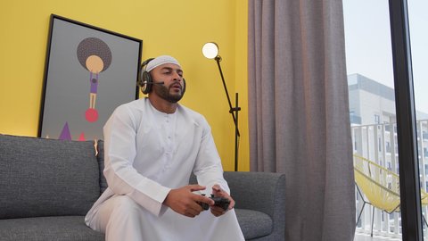 Emirati Arab Gulf man wearing kandura, taqiyah and headphones, holding joystick, entertainment video games, gestures of defeat and anger, expressions of challenge and loss, integrating modern technology in fun and entertainment, spending a good time