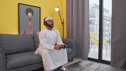 دمج التقنيات الحديثه فى المتعة والتسلية ، قضاء اوقات ممتعه ، رجل عربي اماراتي خليجي يرتدي الكندورة والطاقيه وسماعات الرأس ، يمسك عصا التحكم ، ألعاب الفيديو الترفيهية، ايماءات الهزيمة والغضب