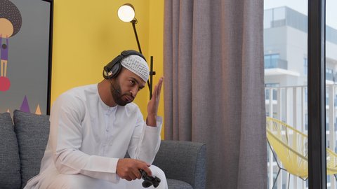 ايماءات الهزيمة والغضب، رجل عربي اماراتي خليجي يرتدي الكندورة والطاقيه وسماعات الرأس ، يمسك عصا التحكم ، ألعاب الفيديو الترفيهية، رفع اليد بتعابير الخسارة، دمج التقنيات الحديثه فى المتعة والتسلية ، قضاء اوقات ممتعه