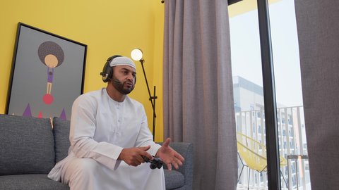 رجل عربي اماراتي خليجي يرتدي الكندورة والطاقيه وسماعات الرأس ، يمسك عصا التحكم ، ألعاب الفيديو الترفيهية، رفع اليد بتعابير الخسارة، دمج التقنيات الحديثه فى المتعة والتسلية ، قضاء اوقات ممتعه