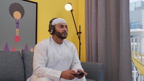 رفع اليد بتعابير الخسارة، دمج التقنيات الحديثه فى المتعة والتسلية ، رجل عربي اماراتي خليجي يرتدي الكندورة والطاقيه وسماعات الرأس ، يمسكان عصا التحكم ، قضاء اوقات ممتعه ، ألعاب الفيديو الترفيهية