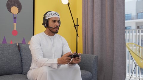 ايماءات التفاعل والتركيز، دمج التقنيات الحديثه فى المتعة والتسلية ، رجل عربي اماراتي خليجي يرتدي الكندورة والطاقيه وسماعات الرأس ، يمسكان عصا التحكم ، قضاء اوقات ممتعه ، ألعاب الفيديو الترفيهية