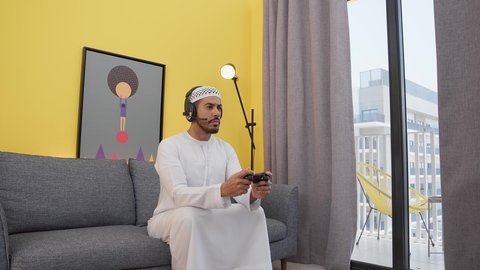 تعابير الاندماج والتركيز ، رجل عربي اماراتي خليجي يرتدي الكندورة والطاقيه وسماعات الرأس ، يمسكان عصا التحكم ، قضاء اوقات ممتعه ، دمج التقنيات الحديثه فى المتعة والتسلية ، ألعاب الفيديو الترفيهية