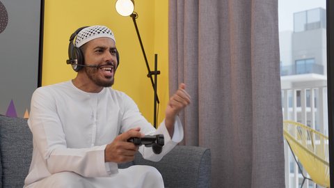 دمج التقنيات الحديثه فى المتعة والتسلية ، رفع قبضة اليد بتعابير الفوز ، رجل عربي اماراتي خليجي يرتدي الكندورة والطاقيه وسماعات الرأس ، يمسكان عصا التحكم ، قضاء اوقات ممتعه ، ألعاب الفيديو الترفيهية