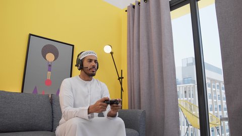 تعابير السرور والسعادة بالانتصار ، ألعاب الفيديو الترفيهية، رجل عربي اماراتي خليجي يرتدي الكندورة والطاقيه وسماعات الرأس ، يمسكان عصا التحكم ، قضاء اوقات ممتعه ، دمج التقنيات الحديثه فى المتعة والتسلية