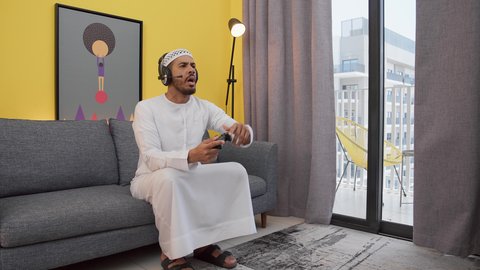 تعابير الغضب والهزيمة والخساره ، ألعاب الفيديو الترفيهية، رجل عربي اماراتي خليجي يرتدي الكندورة والطاقيه وسماعات الرأس ، يمسكان عصا التحكم ، قضاء اوقات ممتعه ، دمج التقنيات الحديثه فى المتعة والتسلية