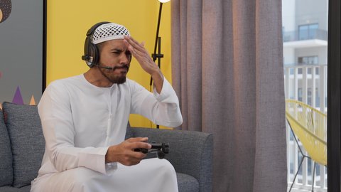 دمج التقنيات الحديثه فى المتعة والتسلية ، تعابير الخساره والهزيمه، ألعاب الفيديو الترفيهية، رجل عربي اماراتي خليجي يرتدي الكندورة والطاقيه وسماعات الرأس ، يمسكان عصا التحكم ، قضاء اوقات ممتعه