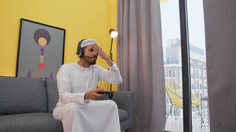 رجل عربي اماراتي خليجي يرتدي الكندورة والطاقيه وسماعات الرأس ، يمسكان عصا التحكم ، دمج التقنيات الحديثه فى المتعة والتسلية ، تعابير الخساره والهزيمه، ألعاب الفيديو الترفيهية، قضاء اوقات ممتعه