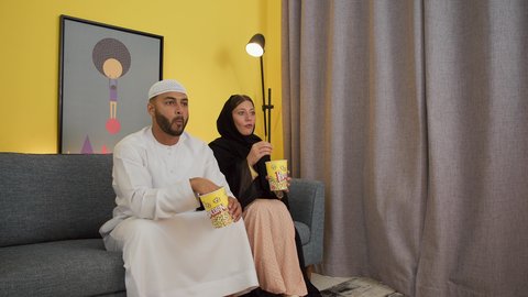 قضاء وقت ممتع بتناول الفوشار اثناء مشاهده التلفاز، مفهوم التسليه والترفيه الجماعي، تصوير لزوجين عربيين خليجيين اماراتيين يجلسان على أريكه مريحه في غرفه المعيشه امام شاشه التلفاز يحملان بيدهما أكواب مليئه بالبوب كورن، تعابير السعاده والسرور