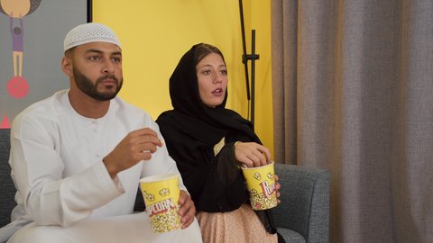 تصوير لزوجين عربيين خليجيين اماراتيين يجلسان على أريكه مريحه في غرفه المعيشه امام شاشه التلفاز يحملان بيدهما أكواب مليئه بالبوب كورن، تعابير السعاده والسرور، قضاء وقت ممتع بتناول الفوشار اثناء مشاهده التلفاز، مفهوم التسليه والترفيه الجماعي