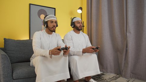الاستمتاع باللعب الجماعي في المنزل، دمج التقنيات الحديثه فى المتعة والتسلية ، اللعب مع الأصدقاء، ايماءات الهزيمة والخسارة، ألعاب الفيديو الترفيهية، شابان عربيان اماراتيان خليجيان يرتديان الكندورة والطاقيه وسماعات الرأس ، يمسكان عصا التحكم