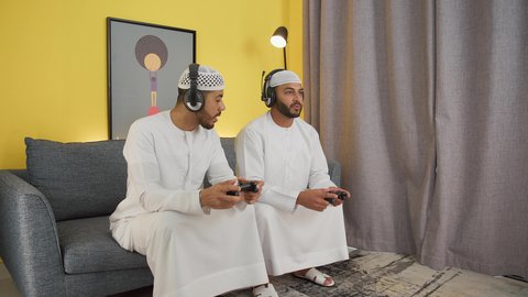 دمج التقنيات الحديثه فى المتعة والتسلية ، اللعب مع الأصدقاء، ألعاب الفيديو الترفيهية، الاستمتاع باللعب الجماعي في المنزل، شابان عربيان اماراتيان خليجيان يرتديان الكندورة والطاقيه وسماعات الرأس ، يمسكان عصا التحكم