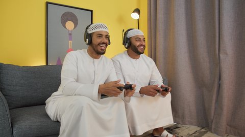 Playing with friends, recreational video games, enjoying group play at home, two young Emirati Gulf Arab men wearing kandora, taqiyah and headphones, holding a joystick, integrating modern technology into fun and entertainment