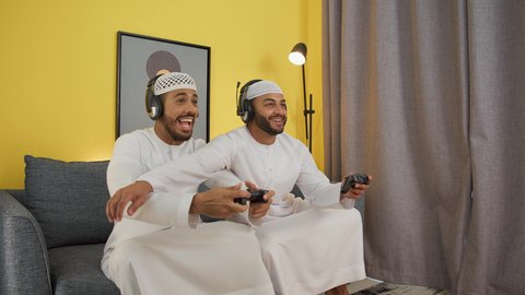 Video games entertainment, playing with friends, enjoying group games at home, two young Emirati Gulf Arab men wearing kandora, taqiyah and headphones, holding a joystick, integrating modern technologies into fun and entertainment
