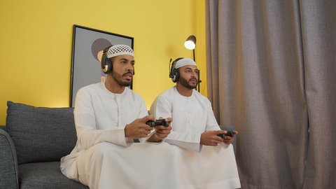 شابان عربيان اماراتيان خليجيان يرتديان الكندورة والطاقيه وسماعات الرأس ، يمسكان عصا التحكم ، دمج التقنيات الحديثه فى المتعة والتسلية ، ألعاب الفيديو الترفيهية، اللعب مع الأصدقاء، الاستمتاع باللعب الجماعي في المنزل
