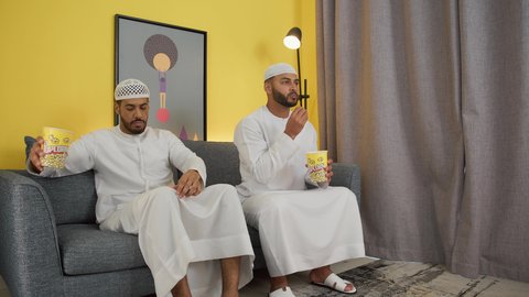 تصوير لصديقين عربين خليجين اماراتيين يرتديان الكندورة والغترة يجلسان على أريكه مريحه في غرفه المعيشه امام شاشه التلفاز يحملان بيدهما أكواب مليئه بالبوب كورن، تعابير السعاده والسرور، قضاء وقت ممتع بتناول الفوشار اثناء مشاهده التلفاز، مفهوم التسليه والترفيه الجماعي