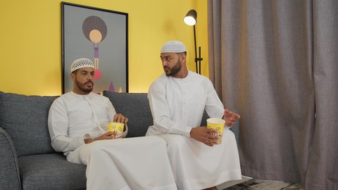 تعابير السعاده والسرور، قضاء وقت ممتع بتناول الفوشار اثناء مشاهده التلفاز، مفهوم التسليه والترفيه الجماعي, تصوير لصديقين عربين خليجين اماراتيين يرتديان الكندورة والغترة يجلسان على أريكه مريحه في غرفه المعيشه امام شاشه التلفاز يحملان بيدهما أكواب مليئه بالبوب كورن