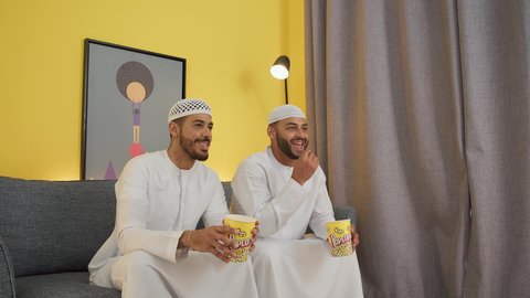 قضاء وقت ممتع بتناول الفوشار اثناء مشاهده التلفاز، مفهوم التسليه والترفيه الجماعي, تصوير لصديقين عربين خليجين اماراتيين يرتديان الكندورة والغترة يجلسان على أريكه مريحه في غرفه المعيشه امام شاشه التلفاز يحملان بيدهما أكواب مليئه بالبوب كورن، تعابير السعاده والسرور