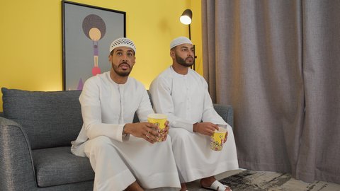 مفهوم التسليه والترفيه الجماعي, قضاء وقت ممتع بتناول الفوشار اثناء مشاهده التلفاز، تصوير لصديقين عربين خليجين اماراتيين يرتديان الكندورة والغترة يجلسان على أريكه مريحه في غرفه المعيشه امام شاشه التلفاز يحملان بيدهما أكواب مليئه بالبوب كورن، تعابير السعاده والسرور