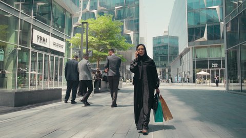 Making phone calls on mobile, photographing a young Emirati Gulf Arab woman wearing an abaya and hijab, carrying shopping bags, buying women's clothes and accessories, modern Gulf women's abayas, a Saudi woman enjoying the pleasure of shopping