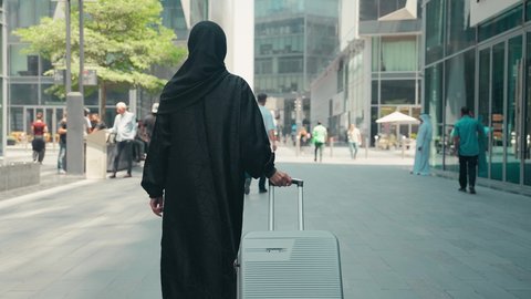 السياحة في إجازة الصيف, قضاء رحلة صيفية ممتعة, سيدة سعودية تستعد لبدء العطلة خارج البلاد , تصوير من الخلف لشابة عربية خليجية اماراتية محجبة ترتدي العباءة تمسك بيدها حقيبة السفر بإيماءات السرور، الاستمتاع بجمال الطبيعة