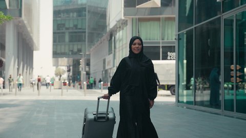 قضاء رحلة صيفية ممتعة, سيدة سعودية تستعد لبدء العطلة خارج البلاد ,السياحة في إجازة الصيف, تصوير لشابة عربية خليجية اماراتية محجبة ترتدي العباءة تمسك بيدها حقيبة السفر بإيماءات السرور، الاستمتاع بجمال الطبيعة