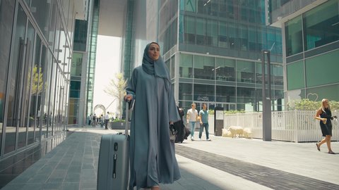 Spending a pleasant summer trip, a Saudi woman preparing to start her vacation abroad, tourism during the summer vacation, a photo of a veiled young Arab Gulf Emirati woman wearing an abaya, holding her travel bag in her hand with gestures of pleasure, enjoying the beauty of nature