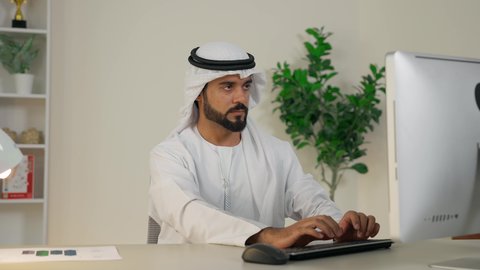 Completing work tasks in the office, integrating work with technology, the development of Emirati companies, managing and organizing business affairs, using a modern and advanced technical device, an Emirati Gulf Arab man wearing a white kandura and ghutra using a computer with gestures of integration and concentration, an office profession and job