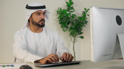 Integrating work with technology, the development of Emirati companies, completing work tasks in the office, managing and organizing business affairs, using a modern and advanced technical device, an Emirati Gulf Arab man wearing a white kandura and ghutra using a computer with gestures of integration and concentration, an office profession and job