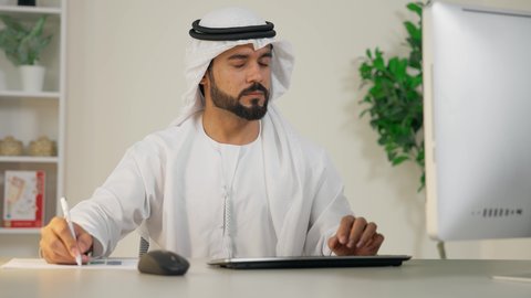 Using a modern and advanced technical device, taking notes on paper, managing and organizing business affairs, completing work tasks in the office, integrating work with technology, the development of Emirati companies, an Arab Gulf Emirati man wearing a kandura and ghutra using a computer, an office profession and job
