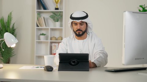 إنجاز مهام العمل، إدارة شؤون الأعمال وتنظيمها عبر الايباد، استخدام جهاز تقني حديث ومتطور، رجل عربي خليجي اماراتي يرتدي الكندورة والغترة البيضاء يجلس خلف الطاولة في المكتب ويستخدم الجهاز اللوحي، مهنة ووظيفة مكتبية، تناول القهوة الساخنة