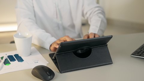 تناول القهوة الساخنة، إنجاز مهام العمل، إدارة شؤون الأعمال عبر الايباد، استخدام جهاز تقني حديث ومتطور، تصوير مقرب لرجل عربي خليجي اماراتي يرتدي الكندورة البيضاء يجلس خلف الطاولة في المكتب ويستخدم الجهاز اللوحي، مهنة ووظيفة مكتبية