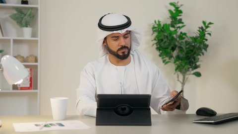 Making a phone call, completing work tasks, managing business affairs via iPad, using a modern and advanced technical device, an Arab Gulf Emirati man wearing a white kandura and ghutra sitting behind a desk in the office and using a tablet, office profession and job, drinking hot coffee