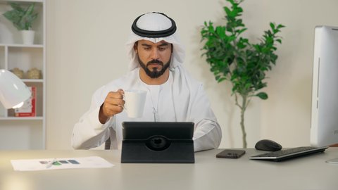 إنجاز مهام العمل، إدارة شؤون الأعمال وتنظيمها عبر الايباد، استخدام جهاز تقني حديث ومتطور، رجل عربي خليجي اماراتي يرتدي الكندورة والغترة البيضاء يجلس خلف الطاولة في المكتب ويستخدم الجهاز اللوحي، مهنة ووظيفة مكتبية، تناول القهوة الساخنة