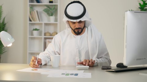 تحليل نتائج البيانات الاحصائية، رجل عربي خليجي اماراتي يرتدي الكندورة والغترة يجلس خلف طاولة المكتب ويعمل على الحاسب المحمول لانجاز المهام، تدوين الملاحظات على الورق، استخدام الالة الحاسبة لاجراء العمليات الحسابية، وظيفة مكتبية في شركات الامارات العربية المتحدة