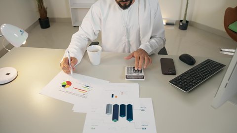 وظيفة مكتبية في شركات الامارات العربية المتحدة، تحليل نتائج البيانات الاحصائية، تصوير علوي لرجل عربي خليجي اماراتي يرتدي الكندورة والغترة يجلس خلف طاولة المكتب يقوم باستخدام الالة الحاسبة لاجراء العمليات الحسابية، تدوين الملاحظات على الورق