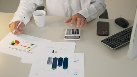 تحليل نتائج البيانات الاحصائية، تدوين الملاحظات على الورق، استخدام الالة الحاسبة لاجراء العمليات الحسابية، وظيفة مكتبية في شركات الامارات، احتساء القهوة في المكتب، تصوير علوي لرجل عربي خليجي اماراتي يرتدي الكندورة يجلس في مكتبه ويقوم بإنجاز مهام العمل