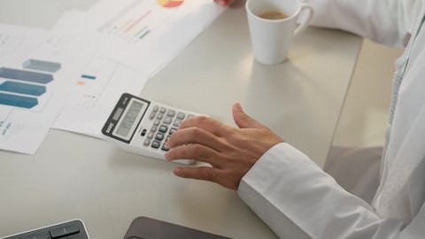 استخدام الالة الحاسبة لاجراء العمليات الحسابية، وظيفة مكتبية في شركات الامارات، تحليل نتائج البيانات الاحصائية، تدوين الملاحظات على الورق، احتساء القهوة في المكتب، تصوير علوي لرجل عربي خليجي اماراتي يرتدي الكندورة يجلس في مكتبه ويقوم بإنجاز مهام العمل